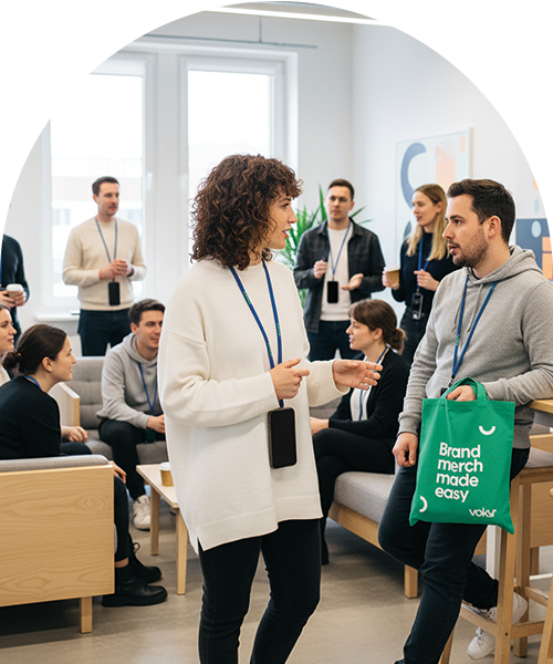 En kvinna i en vit tr&ouml;ja pratar med en man som h&aring;ller i en gr&ouml;n tygkasse med texten "Brand merch made easy", omgiven av andra unga yrkesverksamma i en modern kontorsmilj&ouml;.