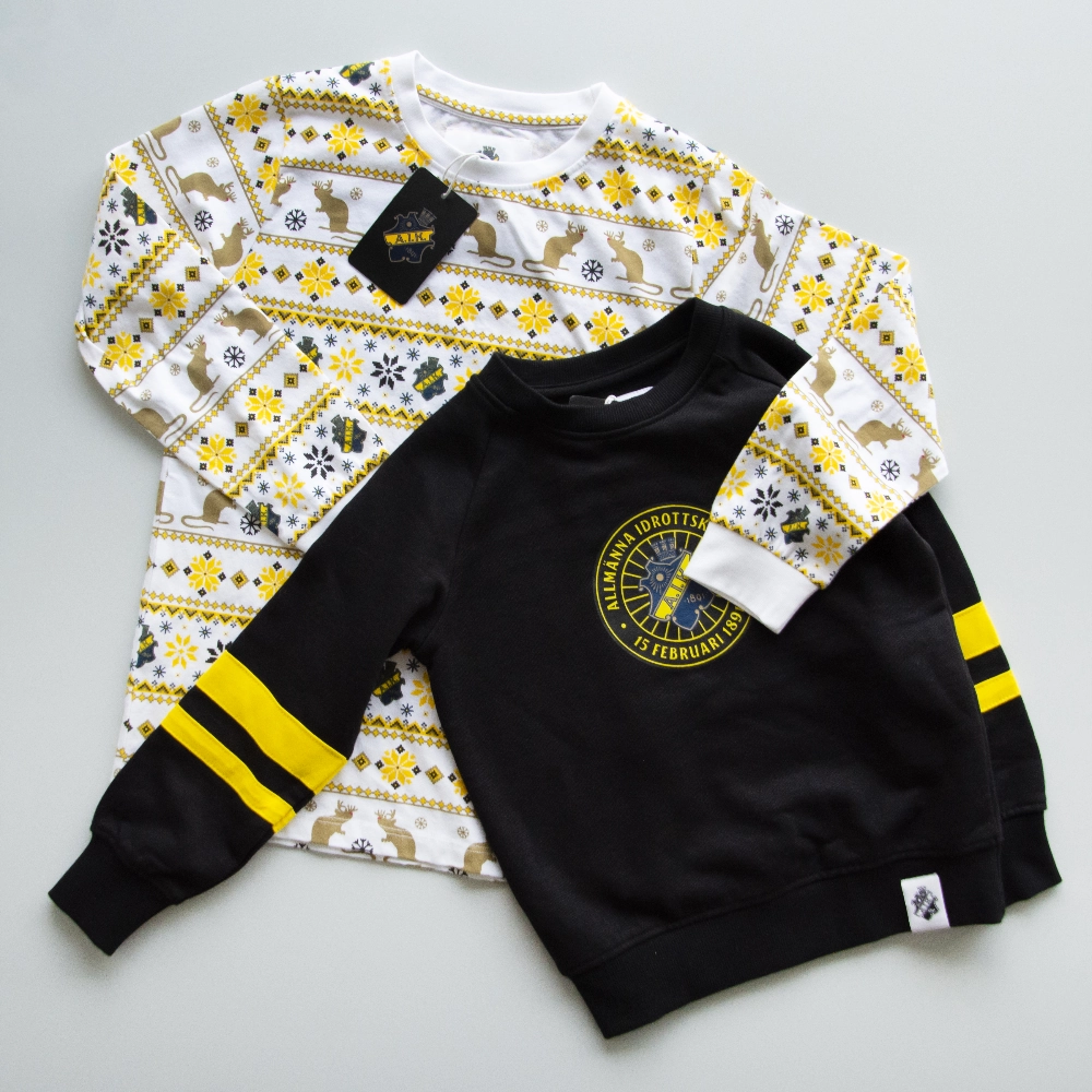  Två AIK barntröjor på ett ljust grått underalag: en vit med ren- och snöflingsmönster i gult och svart, och en svart med gula ärmränder och AIK-klubbmärket på bröstet